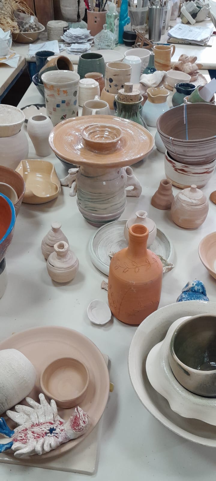 שולחן עבודה בסטודיו עם מגוון רחב של כלי קרמיקה בשלבי יצירה שונים